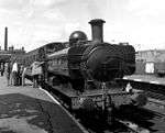 A black and white photo showing a pannier tank locomotive with round windows waiting at a station platform. The following train consists of four passenger coaches, with a tall brick chimney in the background. A group of seven men and boys are looking into the cab of the locomotive. On the side of the tank the letters K W V R can just be made out.