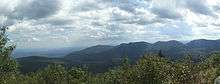 A view over short trees to distant mountains, dappled by intermittent sun and shadow. To the left a river flows through a much lower landscape