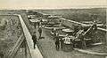 an elevated photo of five guns of three kinds in a row, with several people posed along a fort parapet