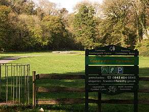 Cairn in the distance in sunshine, with trees in leaf to its left, right and rear. To its front lies flat ground of short grass. An asphalt path leads from the left past the tumulus. The shaded foreground has a kissing gate, a wooden fence and a Forestry Commission welcome sign in Welsh (first) and English.