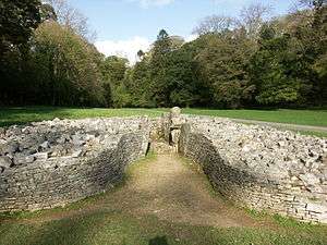 Front view of cairn, its boulders retained by a short, coursed, dry-stone wall that forms a bell-shaped courtyard at its entrance. The cromlech is set in flat ground of short grass. Trees are mainly in leaf to its rear.