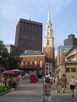 Ground-level view of a brick church with a large, white, tapering spire; a brown skyscraper is visible in the distance, with several shorter high-rises located closer to the church.