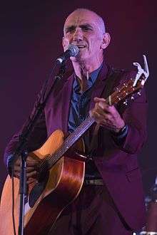 Head and shoulders of a fifty-two-year-old male standing at a microphone. He is shown in right profile, wearing a harmonica in a cradle and staring into the distance.