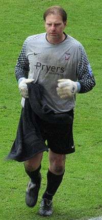 A man with brown hair who is wearing a grey top, black shorts and black socks. He is standing on a grass field.