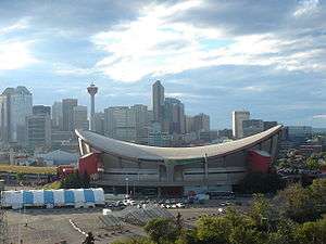 Exterior shot of an indoor arena.  The building has a sloped roof in the shape of a reverse hyperbolic paraboloid and a primarily-concrete outer facing with red towers at the corners.  Several skyscrapers are visible in the background.