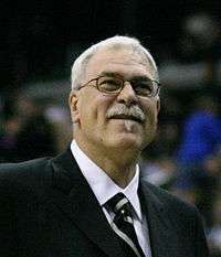 A man with white hair, wearing a black suit, white shirt and tie, at a basketball game.