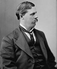 A man with dark hair and a dark mustache wearing a high-collared white shirt, black jacket, and tie