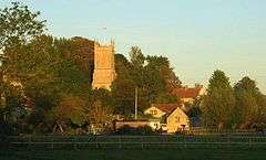 A view across fields to houses and the square tower of a yellow stone church.