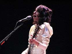 The upper body and face of a female musician. She is wearing a white Victorian-style dress and performing a song on an electric guitar. A microphone is in front of the musician.