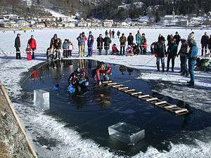  A diver is visible underwater in a hole cut in the ice cover of a small lake. Blocks of ice cut to form the hole are stacked to one side, and a second diver sits on the edge of the hole with his legs in the water. A rough wooden ladder bridges the hole, and a third diver kneels to one side. The dive site is cordoned off with a red and white tape, and other members of the support team stand to the side, with onlookers outside the cordon. There is a residential area along the shoreline in the background.