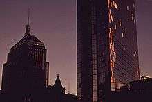 Two dark buildings rise into the early evening sky. The tower on the right is spotted with plywood on its side.