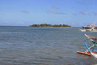 Small low green island in blue sea, at about 600 m, with several palm trees on top.  To right prows of four bangkas - local boats for fishing or diving.