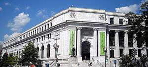 The corner of a white stone, classically designed, 4 story building. The door is on the closer side of the corner, is on the second story with a grand staircase up to it, and is flanked by two ionic columns.