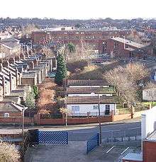 A strip of grass recedes in a straight line into the distance towards a large yellow brick building. Three single storey huts stand on the grass