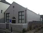 Three-quarter view of a small stuccoed building with a pediment.  The longer side is a blank wall; a flint boundary wall runs alongside it.  The façade has two simple rectangular windows flanking a round-arched entrance, and a small round window below the pediment.