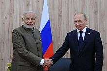 Two standing men are pictured shaking hands. The first is dressed in Indian clothing; the second is in a Western business suit; both standing behind a Russian flag.