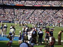 Full stands, both teams on the field, cheerleaders and lots of people milling around