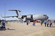 Colour photo of a grey military aircraft on the tarmac of an airport with people in casual clothes walking in front of it