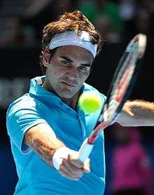 A brown-haired man in a light blue polo shirt and both white wristband and bandanna with a light blue nike logo, who is hitting a backhand with the tennis ball in the foreground