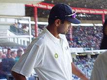 A male, dark-skinned cricketer in a white short-sleeved shirt and dark blue baseball cap with sunglasses on the peak of the cap. He is standing high up in a cricket stand with a large number of spectators in the background.