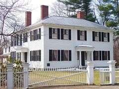 A white two story house, whose main section is square, although there is a wing extending off the back. There is a white fence with a gate across a gravel drive.