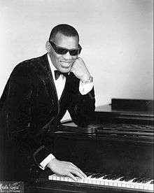 Ray Charles, wearing sunglasses, leaning on a piano from the side with his hand reaching towards the keys.