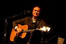 A man sits on a stool, smiling and facing the camera while playing guitar. He wears brown, and the background behind him is black.