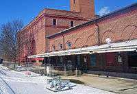 A brick building with snow-covered ground and a bare tree at the lower left. The portion of the building nearer the camera, on the right, has large rounded indentations in it and a metal roof hung from its side by cables over a bare concrete surface. The portion on the left, farther away, has been fenced off. It is taller, with a large square section rising to the top of the image in the center.