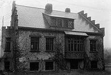 A black and white image of the house, seen from the front on a wet day, with stepped gables and a shed dormer. It is covered by ivy (not in bloom) and the chimneys have Gothic-arch decor