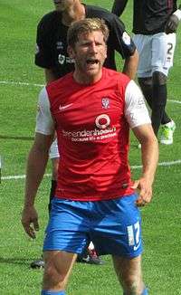 A man with short brown hair is wearing a red top, blue shorts and white socks. He is standing on a grass field.