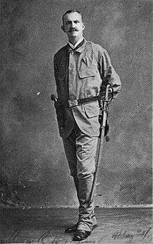 A monochrome photograph portrait of a tall, thin man with a moustache and thinning hair, wearing a plain utilitarian uniform with gaiters and a wide belt holding a long, curved sword and holster with handgun