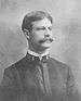 Head and shoulders of a white man with a wide mustache and carefully combed hair wearing a plain military jacket with a single bar on each side of the collar.