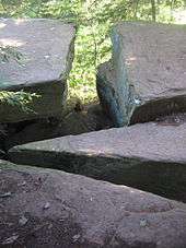 Photo of four large flat-topped boulders divided by narrow splits. Green foliage is visible at the top of the image creating a t-shape in the rocks