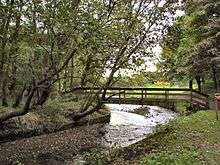 View down a small river, bending to the left, crossed by a wooden footbridge in the middle distance.