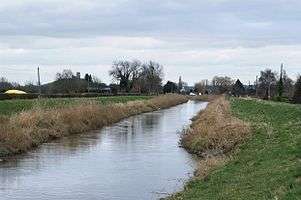 Fairly straight stretch of water between grassy banks. In the distance are buildings and trees with a church on a hill.