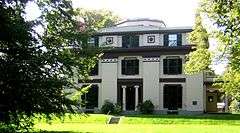 A white house with dark trim, including decorative elements. The house has three stories, and there is an octagonal element protruding above the top floor.