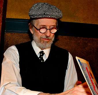 An elderly man with a white beard, round glasses, a beret-like hat, a dark vest, and a necktie.  He faces down right, looking into an open book.