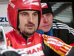 A man with a goatee wears a red-white-and-yellow tight jumpsuit, with a red-and-white vest over it, and a red helmet with a raised full-faced visor. He shows a concentrated look. Partially hidden behind him is another man, wearing a black tracksuit jacket and winter cap.