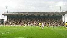 An impressive grandstand behind a goal net, filled with people, viewed from the other end of the stadium.