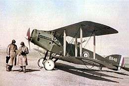 Two men in long leather coats and leather helmets stand in front of a biplane in RAF colours.