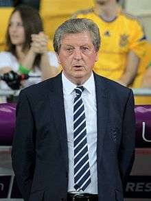 A photograph of a grey-haired, middle-aged man at a football match. He is wearing a black suit, a white shirt and a black and white striped tie. He is watching the game from the sideline.