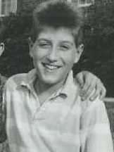 A back and white photo of a Caucasian teenaged male. He is wearing a light colored polo with stripe's, button's by the neckline and a neck collar. Another person's ear can be seen with his arm on the shoulder of the teenage male.