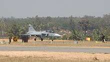 Jet aircraft in the distance preparing to take off from rural airport surrounded by green trees