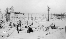 A snowy hill with stone barriers and barbed wire in front, and a distant bunker in behind.