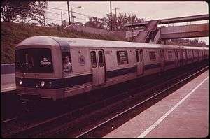 A Staten Island Railway train composed R44 subway cars on the Staten Island Railway. This image, taken in 1973, shows the cars with a since-removed blue stripe toward the bottom of the car body. The train is arriving at a platform to the left; the photo is taken from another platform to the right and in the foreground. The station is in a right-of-way below street level, and a covered footbridge connecting the two platforms is located to the right.