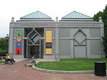 A one story symmetrical granite building with two green peaked roofs either side of center. The entrance door is on the left and a similar design is on the right made out of a slightly darker granite.