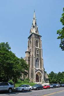 A tall brownish stone church steeple, intricately detailed, rising high over a street with cars parked on it. A grassy area with trees is on the left.