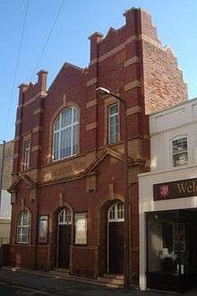 A tall building with deep red brickwork and yellowish stone dressings. There are two identical entrances: wooden doors in recessed stone doorways with prominent yellow keystones.  A window to their left is in a similar recess.  Two pediments flank a stone frieze with the words "THE SALVATION ARMY".  The upper floor has two rectangular windows and a central arched window with another keystone.