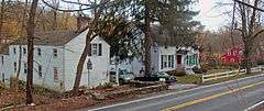 Three houses seen from an elevated position across a road. The two on the left are white, with the leftmost facing the road side on. Between it and the house next to it is a driveway with two cars and a tall evergreen tree. Further down the road, at the right, the house is red.