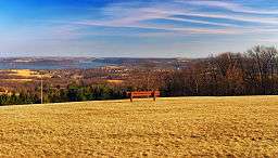 A red bench on a large expanse of brown cut grass, overlooking rolling forested hills and a lake